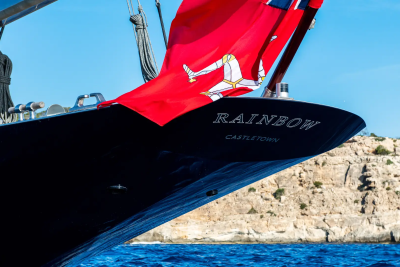 A dark hull of a yacht named "Rainbow" flies a red flag with a three-legged symbol, set against a backdrop of a rocky coastline and blue water.