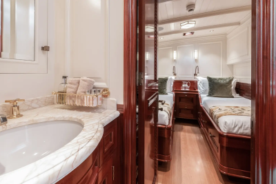 A marble-topped vanity with a sink is in the foreground, while a mirrored hallway leads to a bedroom with two beds in the background.