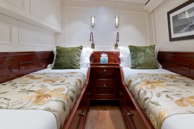 Two twin beds with floral comforters and green pillows are in the foreground, while the background features a nightstand with lamps and a framed picture on the wall.