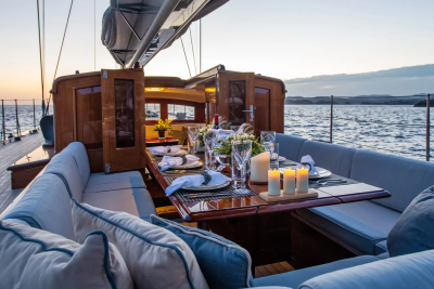 A beautifully set dining table on a yacht's deck is bathed in the soft light of a sunset, with the calm sea and distant coastline forming the backdrop.