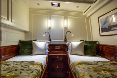 Two neatly made beds with plush pillows and patterned comforters are flanked by dark wood nightstands, while a framed nautical painting hangs on the paneled wall in the background.