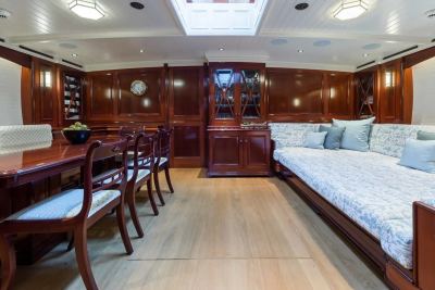 A polished wooden dining table with chairs in the foreground is set against a backdrop of a luxurious yacht interior with a long sofa and ornate cabinetry.