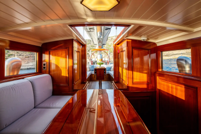A polished wooden interior of a yacht with a plush sofa in the foreground and a view of the sea and rocky shore through open doors in the background.