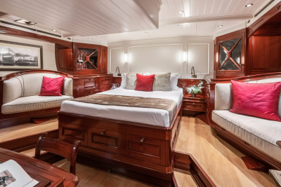 A luxurious yacht cabin features a central bed with plush pillows, flanked by ornate wooden seating and cabinetry, with a framed picture on the wall in the background.
