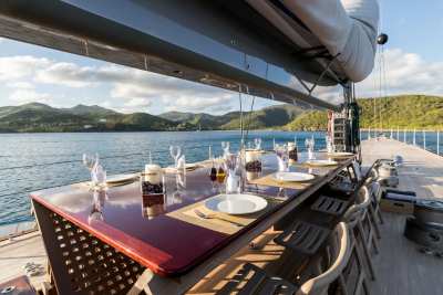 A long table set for dinner on a yacht's deck overlooks a calm sea and lush green hills under a bright sky.