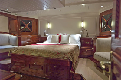 A luxurious yacht cabin features a large bed with white linens and red accents in the foreground, while the background reveals polished wood cabinetry and soft lighting.