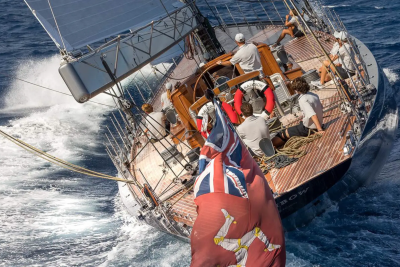 A large sailboat with a crew on deck sails through choppy blue water, with a flag prominently displayed in the foreground.
