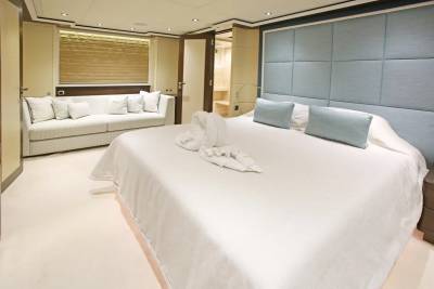A large white bed with swan-shaped towels in the foreground is in a luxurious bedroom with a light blue tufted headboard, while a white sofa and an open doorway leading to another room are in the background.