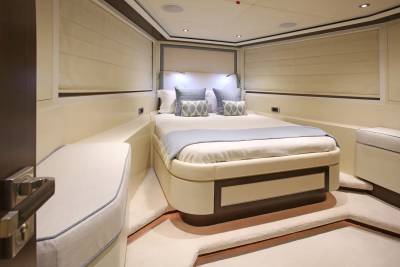 A luxurious yacht bedroom with a plush bed in the center and a door handle in the foreground, set against a backdrop of cream-colored walls and blinds.