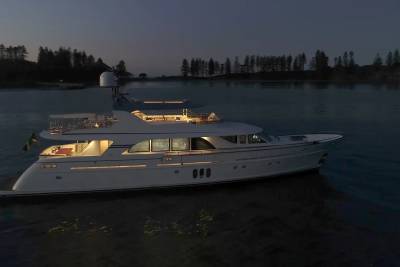 A luxurious white yacht with illuminated windows floats on calm water in the foreground, while a dark, tree-lined shore is visible in the dusky background.