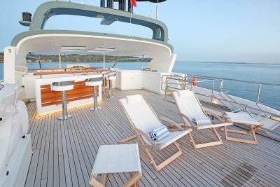 Deck chairs and a bar are set up on a yacht's wooden deck, with a calm ocean and distant coastline in the background.