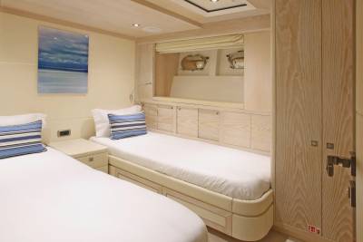 Two neatly made twin beds with blue and white striped pillows are in the foreground, while the background features a wooden closet and a framed seascape artwork.