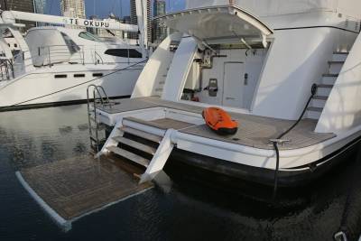 A white yacht with a lowered swim platform and an orange rescue device in the foreground is docked next to another white yacht with city buildings in the background.