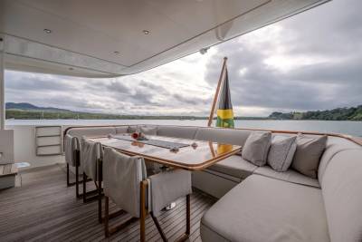 A spacious outdoor dining area with a large U-shaped sofa and chairs in the foreground overlooks a calm body of water with a flag and distant tree-lined hills under a cloudy sky in the background.