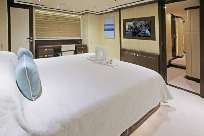 A large white bed with blue pillows dominates the foreground, while the background reveals a modern bedroom with a desk, television, and an open doorway leading to another room.