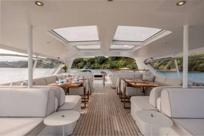 Luxurious white sofas and dining tables fill the foreground of a yacht's deck, while the background reveals a tranquil bay with lush green hills.