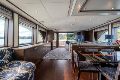 A luxurious yacht interior features a patterned sofa in the foreground and a dining area leading to an open deck with a water view in the background.