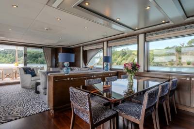 A dining table with floral centerpiece sits in the foreground of a luxurious yacht's interior, with large windows revealing a scenic waterfront view in the background.