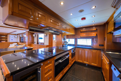 A modern yacht galley with a sleek black countertop and wooden cabinets in the foreground, and a dining area with white furniture visible in the background.