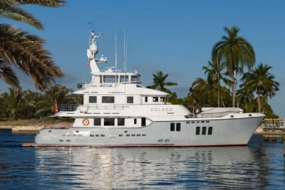 A large white yacht named "SOLACE" floats on calm blue water in the foreground, with lush green palm trees and a clear blue sky forming the background.
