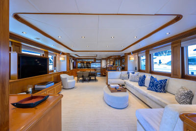 A spacious yacht interior features a large sectional sofa with blue-accented pillows in the foreground, while the background reveals a dining area and windows overlooking the water.