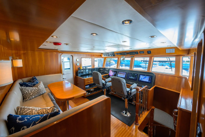 A comfortable seating area with a table is in the foreground, while the background features a ship's bridge with navigation equipment and windows overlooking a marina.