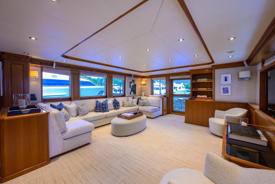 A plush sectional sofa with blue and white patterned pillows faces an oval ottoman in the foreground, while large windows in the background reveal a glimpse of a docked yacht.