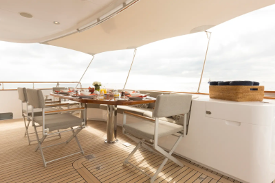 A dining table set for a meal on a yacht's deck is in the foreground, with the ocean and a cloudy sky visible in the background.