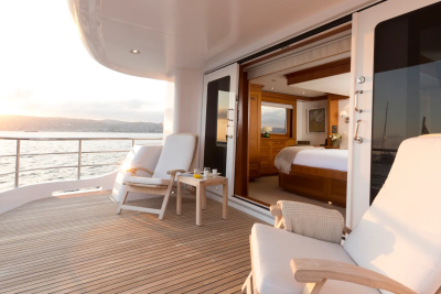 Two white lounge chairs and a small table with drinks sit on a wooden deck in the foreground, while an open doorway reveals a luxurious bedroom with a large bed and wooden accents in the background, all set against a backdrop of calm water and a distant coastline bathed in soft sunlight.