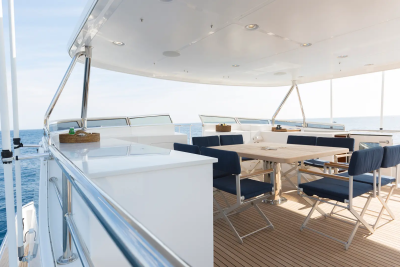 A white yacht's deck with a dining table and chairs in the foreground is set against a backdrop of the calm blue sea and a distant ship.