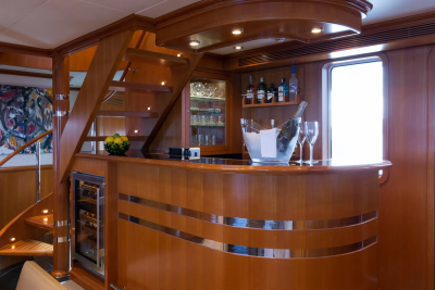 A polished wooden bar with an ice bucket and champagne sits in the foreground, while a spiral staircase and a colorful abstract painting adorn the background of this yacht's interior.