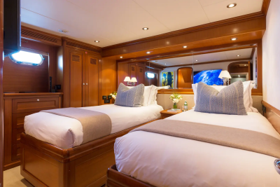 Two neatly made twin beds with gray throws and patterned pillows fill the foreground, while a mirrored wall reflecting a television and a porthole window adorns the background of this luxurious cabin.
