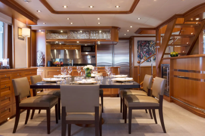 A formal dining table is set for a meal in a luxurious yacht's dining room, with a well-equipped galley and a staircase visible in the background.