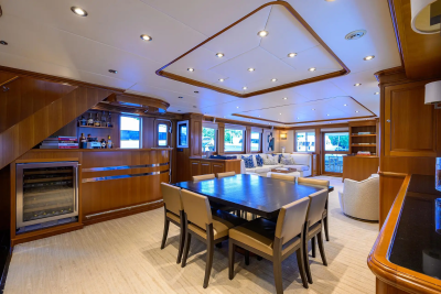 A polished wooden dining table with chairs in the foreground is set within a spacious, well-lit yacht interior featuring a bar and lounge area in the background.