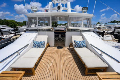 Two white lounge chairs with blue and white patterned pillows sit on a teak deck in the foreground, while a white yacht with a covered seating area and other boats docked in the background are visible under a bright blue sky.