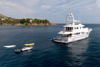 A large white yacht floats on calm blue water in the foreground, with smaller boats and a yellow kayak nearby, while a tree-covered hillside with houses forms the background.