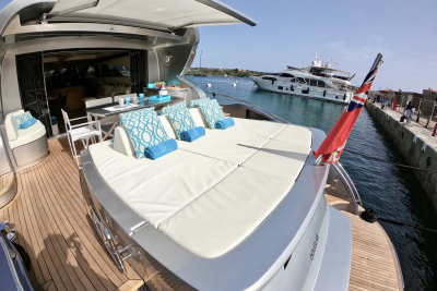 A luxurious yacht with a sunbathing deck in the foreground is docked next to another large yacht in the background, with a clear blue sky overhead.