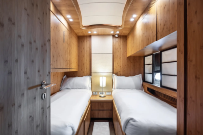 Two neatly made twin beds with white linens flank a nightstand with a lamp in the foreground, while the background features warm, wood-paneled walls and a ceiling with recessed lighting.