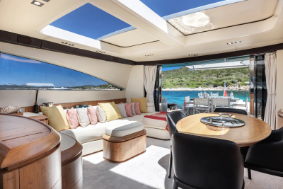 A luxurious yacht interior with a plush sofa and dining table in the foreground, opening to a scenic view of a sun-drenched coastline and turquoise waters in the background.