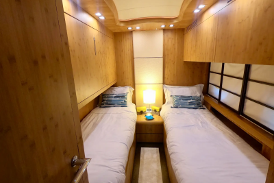Two neatly made twin beds with blue and white striped pillows flank a nightstand with a lamp in the foreground, while the background features wood-paneled walls and cabinets with soft lighting.