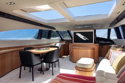 A modern yacht interior features a dining table with black chairs in the foreground and a large television screen and comfortable seating in the background, with a scenic ocean view through the windows.