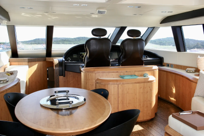 A circular wooden dining table with black chairs sits in the foreground, while the background features a yacht's helm with two leather seats and large windows overlooking a sunny, coastal landscape.