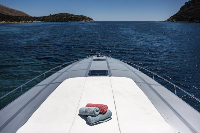 A sleek yacht with folded towels on its deck glides through calm blue water towards a distant, hilly coastline under a clear sky.