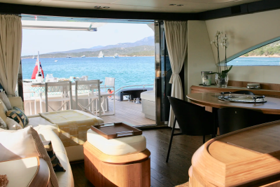 A luxurious yacht interior with a comfortable seating area in the foreground opens to a sun-drenched deck with a dining table and a stunning view of the turquoise sea and distant sailboats in the background.