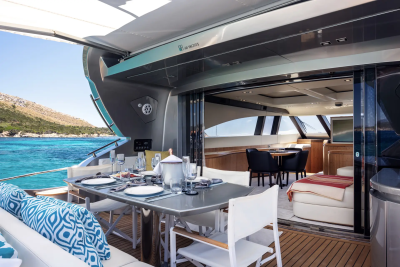 A beautifully set dining table on a yacht's deck overlooks a turquoise sea and a hilly coastline.