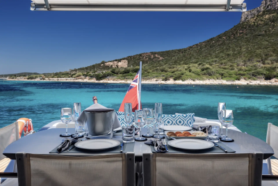A set dining table on a yacht's deck overlooks a tranquil turquoise sea and a lush, mountainous coastline under a clear blue sky.