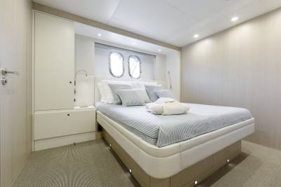 A neatly made bed with striped linens and plush pillows sits in the foreground, while the background features two porthole windows and a wall with textured wallpaper.