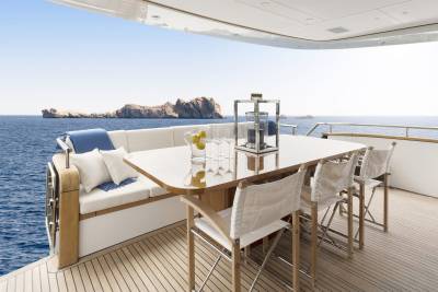 A white outdoor dining set with a pitcher of lemons and glasses sits on a yacht's deck, with a rocky island and blue ocean in the background.