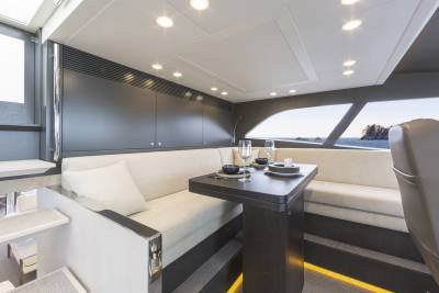 A modern yacht interior features a plush L-shaped sofa and dining table in the foreground, with a scenic ocean view visible through large windows in the background.