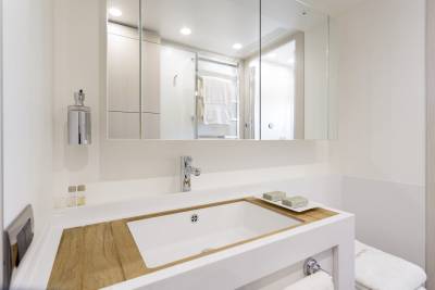 A modern white bathroom sink with a wooden countertop is in the foreground, while a large mirror reflecting a towel rack and shower is in the background.